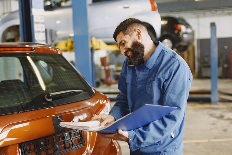 Mecânico de macacão azul sorrindo ao lado de um carro laranja