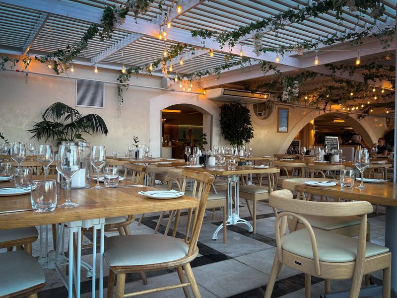 Salão do restaurante com mesas de madeira, cadeiras claras, taças e plantas